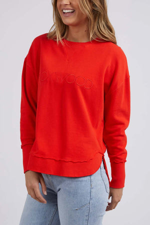 A person in a bright red Foxwood Simplified Crew sweatshirt, renowned for its relaxed fit and 100% cotton fabric, with "OXWOOD" embossed on the front, paired with light blue jeans, smiles while standing against a plain background.