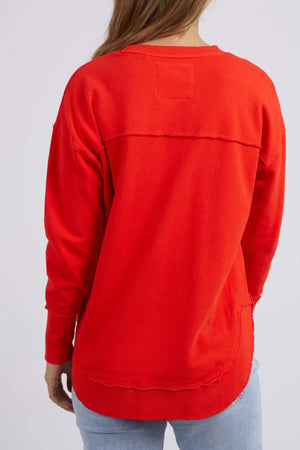 A person wears a bright red Foxwood Simplified Crew sweatshirt with a relaxed fit and rectangular stitch at the neckline. Made from 100% cotton with a curved hem, it's paired with light blue jeans against a plain gray background.