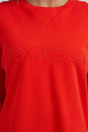Close-up of a person wearing the relaxed fit Foxwood Simplified Crew, a bright red 100% cotton sweatshirt with "FOXWOOD" embroidered in red across the front and a triangular ribbed detail at the neckline.