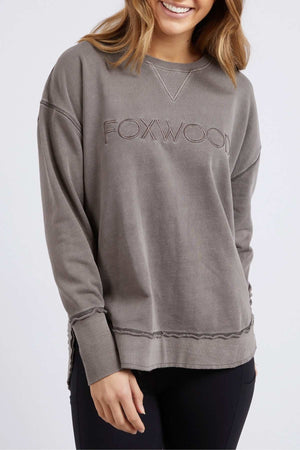 A woman wearing a loose-fitting, long-sleeve, gray Foxwood Simplified Crew with front embroidery smiles while standing against a plain light background.
