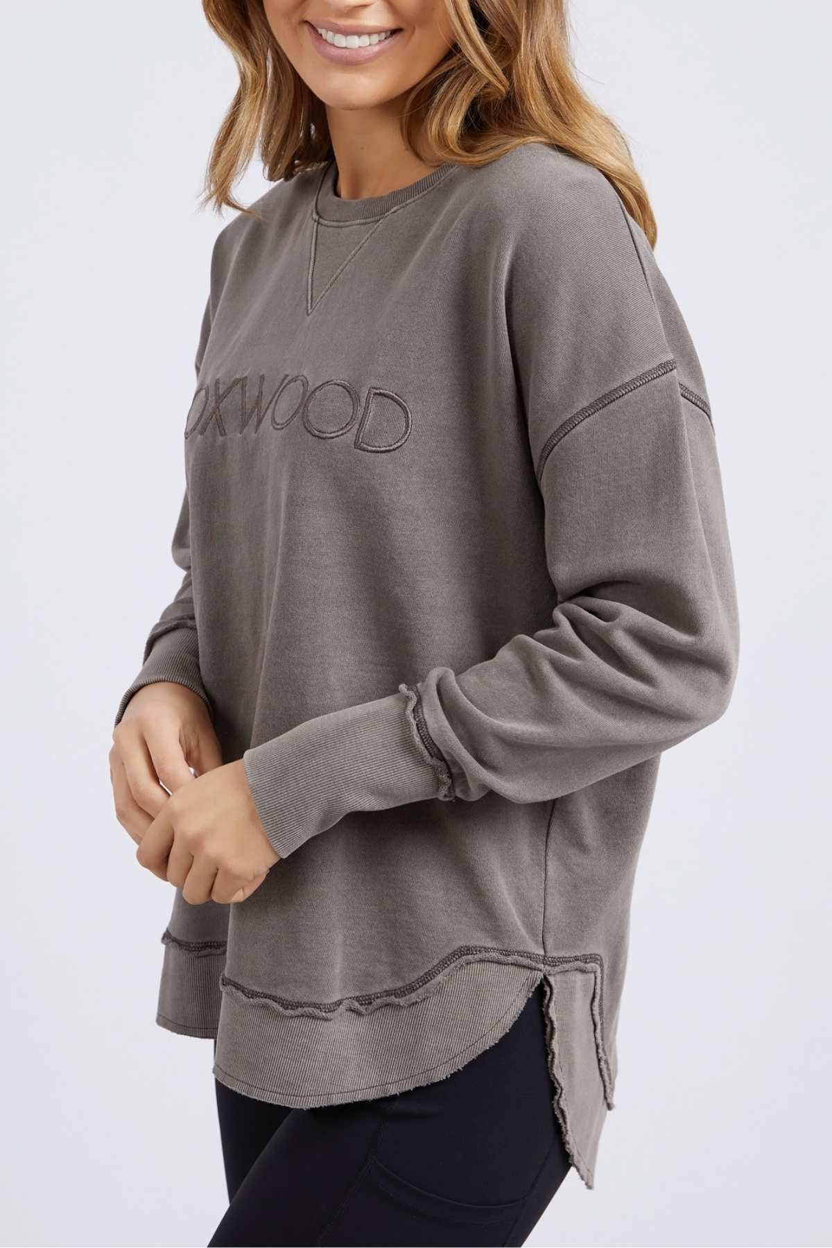 A woman wearing a loose, long-sleeved gray Foxwood Simplified Crew with "OAKWOOD" embroidery on the front and black leggings smiles while standing against a plain light background.