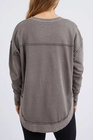 A person with long hair wears the Foxwood Simplified Crew, a relaxed-fit 100% cotton gray sweatshirt with exposed seams, paired with black leggings. They stand with their back to the camera against a plain white background.