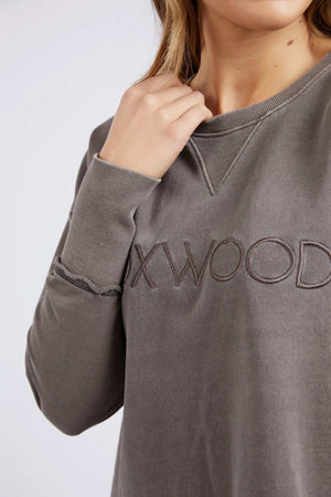 A person wears a grey Foxwood Simplified Crew sweatshirt with "OXWOOD" embroidery across the chest, made from 100% cotton, holding the collar with one hand. The person's face is partially out of frame.