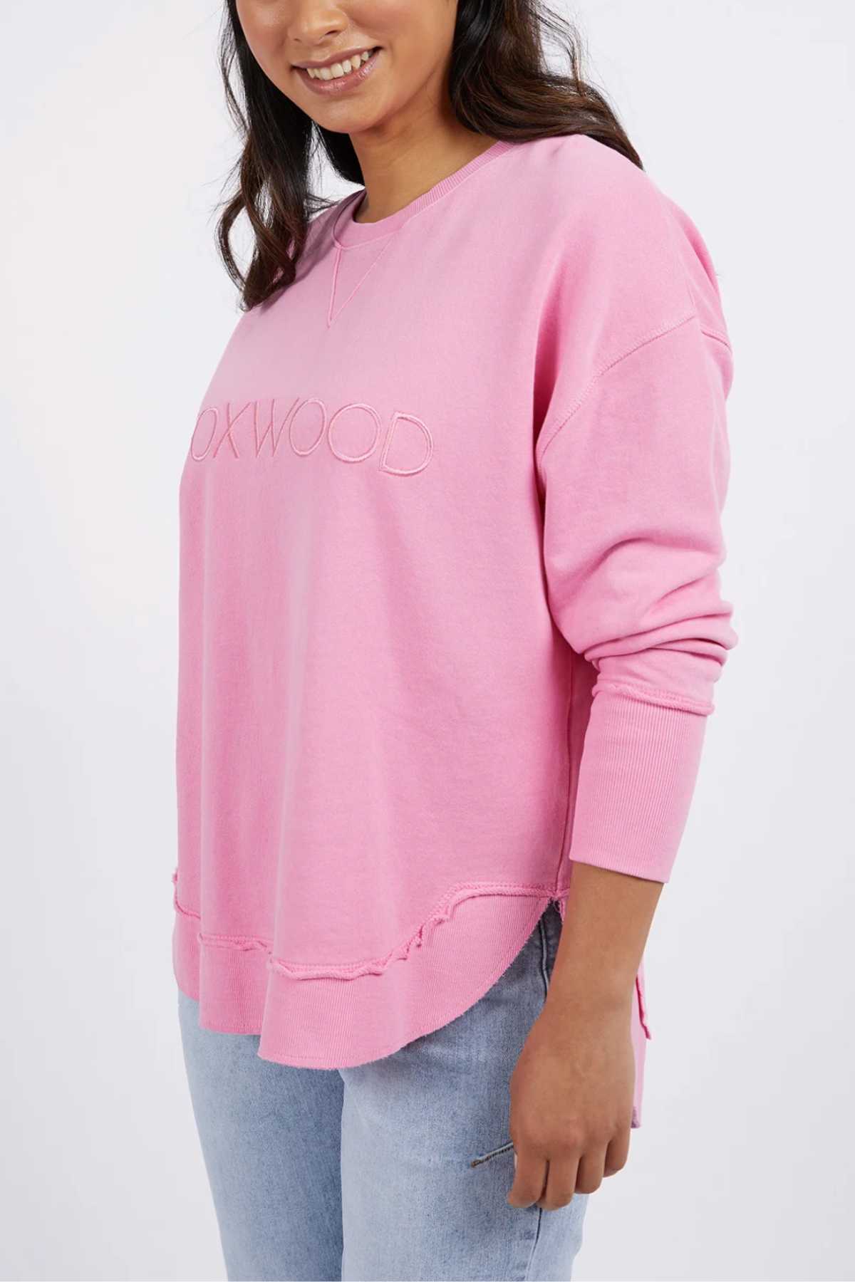 A woman smiles in a loose, pink Foxwood Simplified Crew with embroidered logo and light blue jeans. Made from 100% cotton, she stands against a plain, light background.