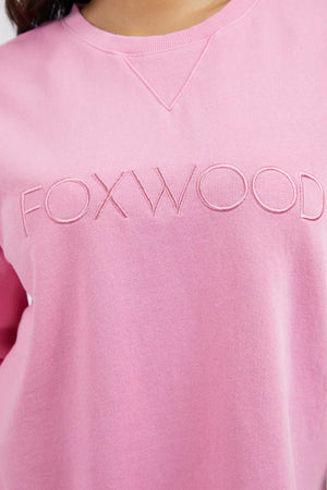 Someone wears the Foxwood Simplified Crew, a light pink 100% cotton sweatshirt with "FOXWOOD" embroidered across the chest in matching pink thread.