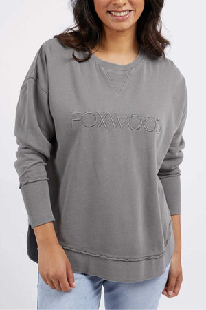 A person wearing a grey Foxwood Simplified Crew sweatshirt with "FOXWOOD" embroidery on the front. They have long, dark hair and are smiling. The person is also wearing light blue jeans and is standing against a plain white background.
