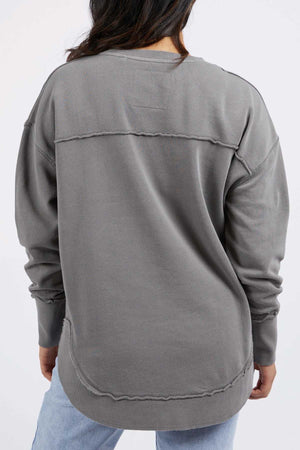 A person with long dark hair is seen from behind wearing a loose-fitting, grey Foxwood Simplified Crew with visible raw-edged seams and distressed details, paired with light-wash jeans. The background is plain white.