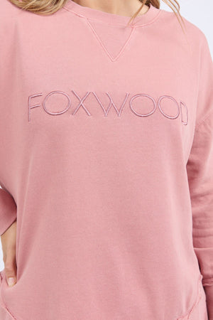 A person wears a relaxed fit, light pink Foxwood Simplified Crew, made from 100% cotton, with "FOXWOOD" embroidered in large capital letters across the chest. Only the torso and part of the arm are visible.