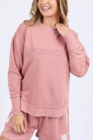 A woman wears the Foxwood Simplified Crew, a relaxed 100% cotton dusty pink sweater featuring "FOXWOOD" embroidery, styled with matching shorts as she smiles and softly holds the collar.