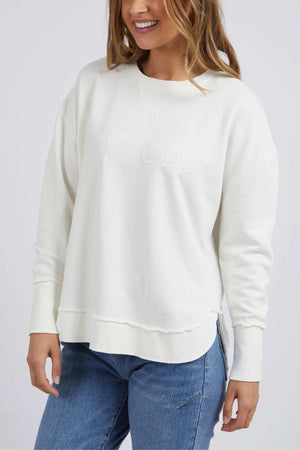 A person wearing the Foxwood Simplified Crew, a white sweatshirt with "Foxwood" subtly embroidered on the front, paired with blue jeans. The individual has long hair and is smiling against a plain white background.