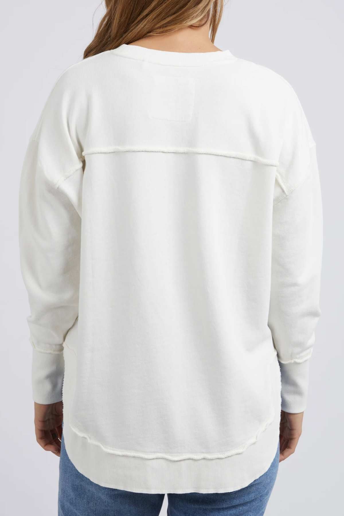 A person with long hair is shown from the back, wearing a Foxwood Simplified Crew in white. This long-sleeved shirt features a distinctive horizontal seam across the upper back and another along the hem. The shirt is paired with blue jeans, set against a light background.
