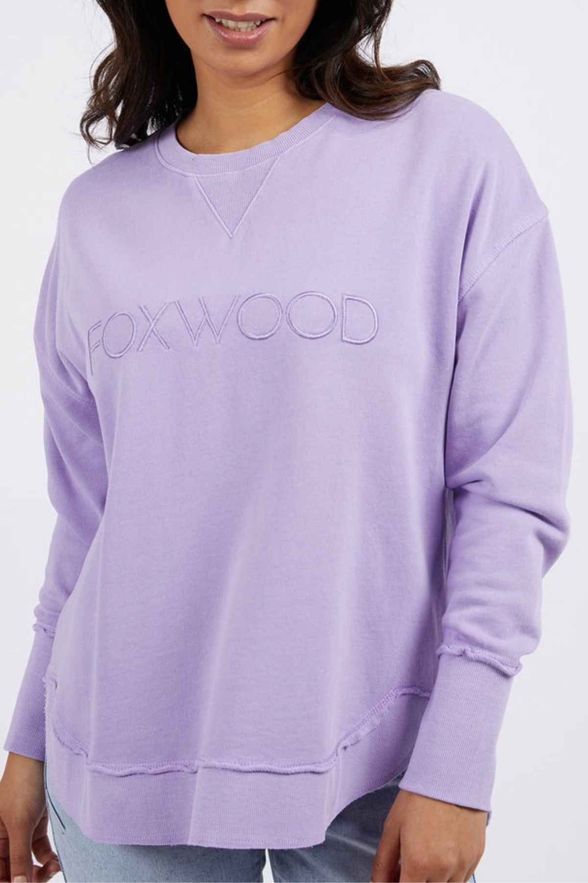 A woman wears a relaxed fit, light purple Foxwood Simplified Crew (100% cotton) with "FOXWOOD" embroidery, paired with light blue jeans. The photo is cropped from shoulders to mid-thigh.