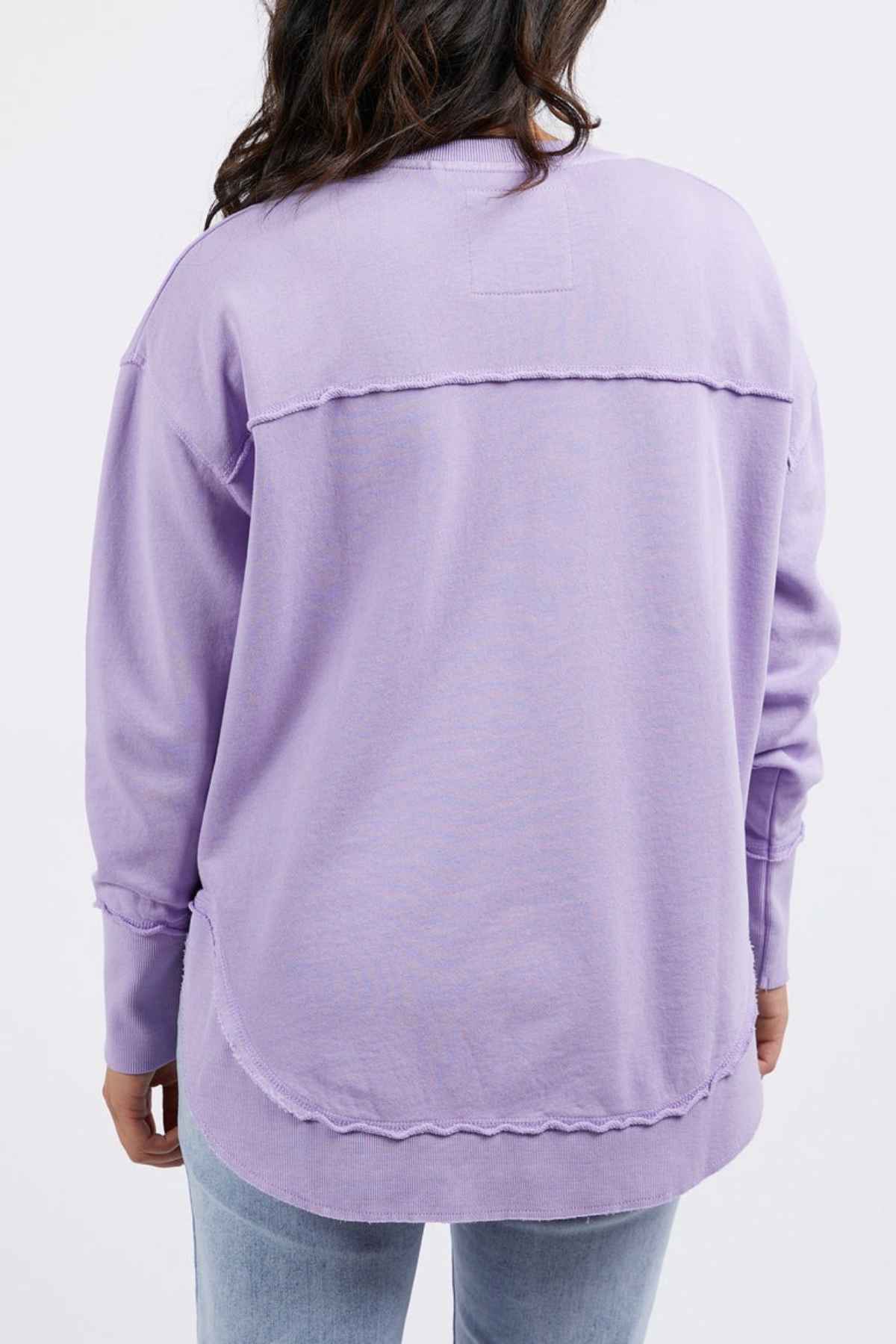 A person with long dark hair is shown from the back, wearing a relaxed fit 100% cotton Foxwood Simplified Crew in lavender, paired with light blue jeans. The background is plain and light colored.