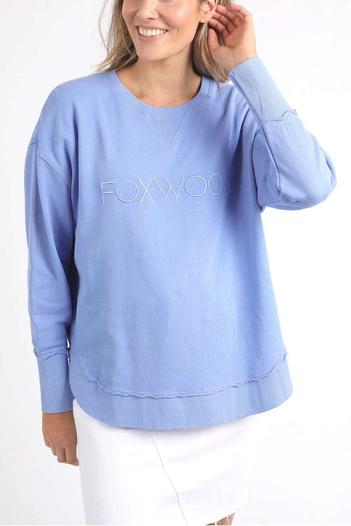 A woman is wearing a Foxwood Simplified Crew sweatshirt in light blue, featuring "FOXWOOD" embroidered on the front. Made from 100% cotton for comfort and breathability, she pairs it with a white skirt. Smiling, she holds her long blonde hair back with one hand against a plain white background.