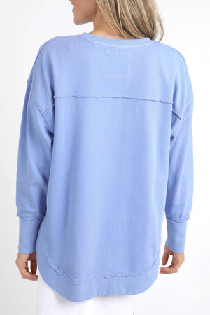A person with long blonde hair is shown from the back, wearing a light blue, long-sleeved Foxwood Simplified Crew sweatshirt made from 100% cotton for comfort and breathability. They are also wearing white pants against a plain white background.