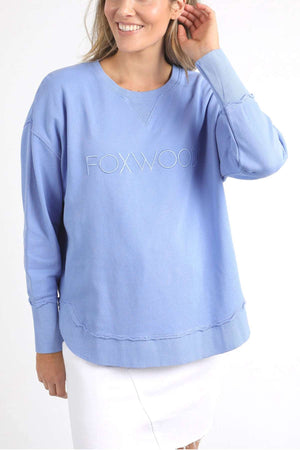 A woman is wearing a Foxwood Simplified Crew sweatshirt in light blue, featuring "FOXWOOD" embroidered on the front. Made from 100% cotton for comfort and breathability, she pairs it with a white skirt. Smiling, she holds her long blonde hair back with one hand against a plain white background.