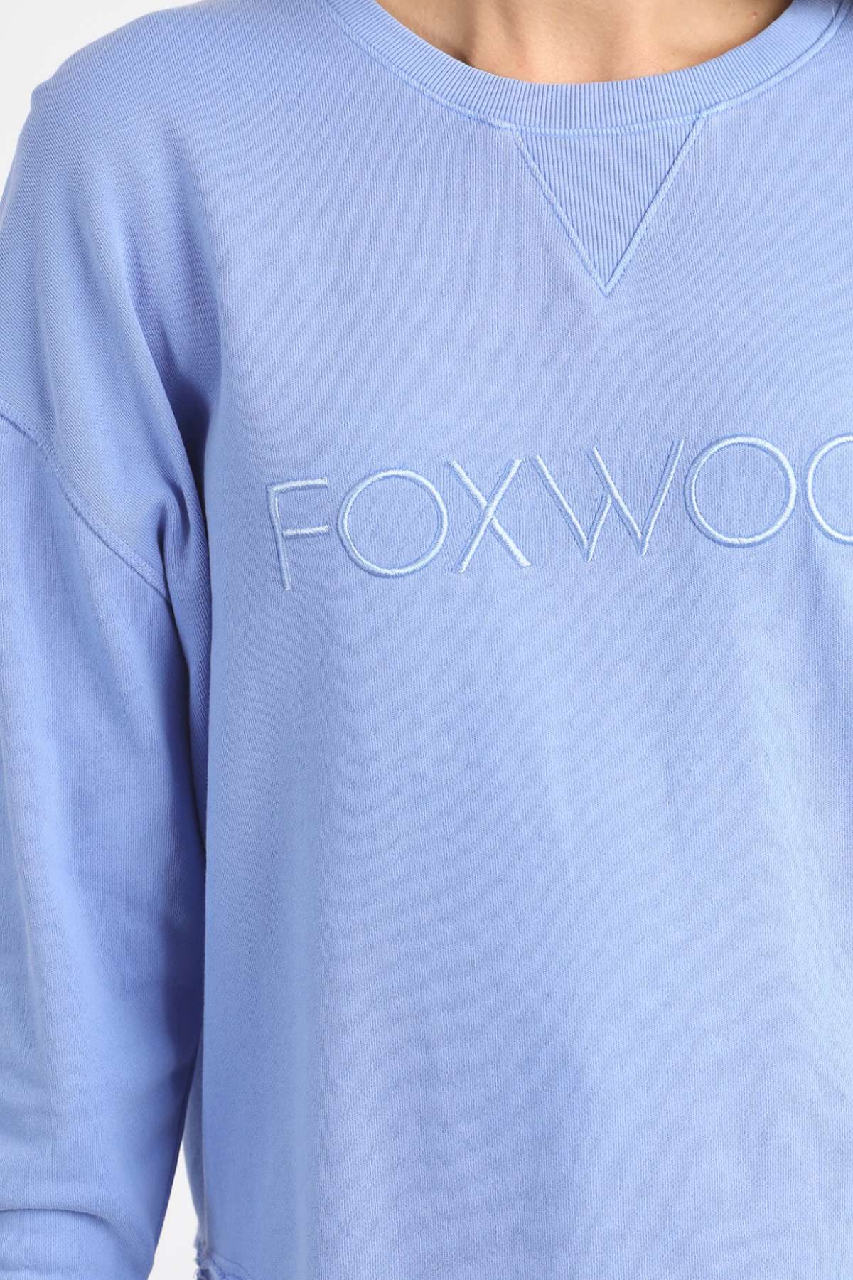 A close-up shows a person wearing the Foxwood Simplified Crew, a light blue sweatshirt crafted from 100% cotton for superior comfort and breathability. The word "FOXWOOD" is embroidered in white across the chest, and the sweatshirt features a ribbed neckline with a distinctive V-shaped stitch detail below the collar.