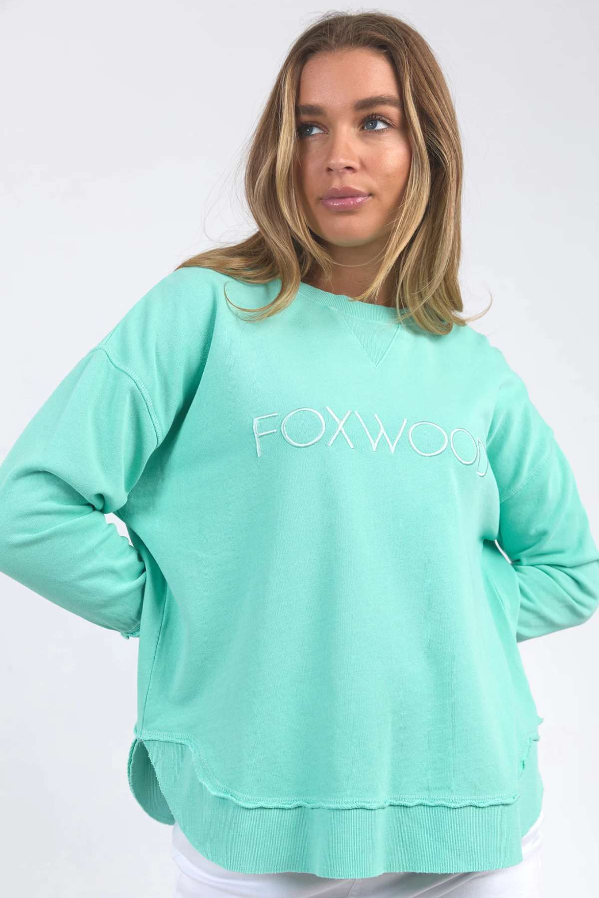 A person with long hair is wearing a mint green Foxwood Simplified Crew, which features "FOXWOOD" embroidered on the front. Crafted from 100% cotton, the sweatshirt offers a relaxed fit. They have their hands partially tucked into the sweatshirt's pockets and are looking slightly to the side against a plain, light-colored background.
