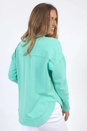 A woman with long, light brown hair smiles while looking over her shoulder. She is wearing a relaxed-fit Foxwood Simplified Crew sweater in mint green, crafted from 100% cotton, paired with white pants. The background is plain white.