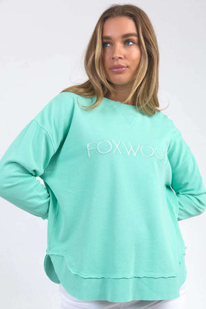 A person with long hair is wearing a mint green Foxwood Simplified Crew, which features "FOXWOOD" embroidered on the front. Crafted from 100% cotton, the sweatshirt offers a relaxed fit. They have their hands partially tucked into the sweatshirt's pockets and are looking slightly to the side against a plain, light-colored background.