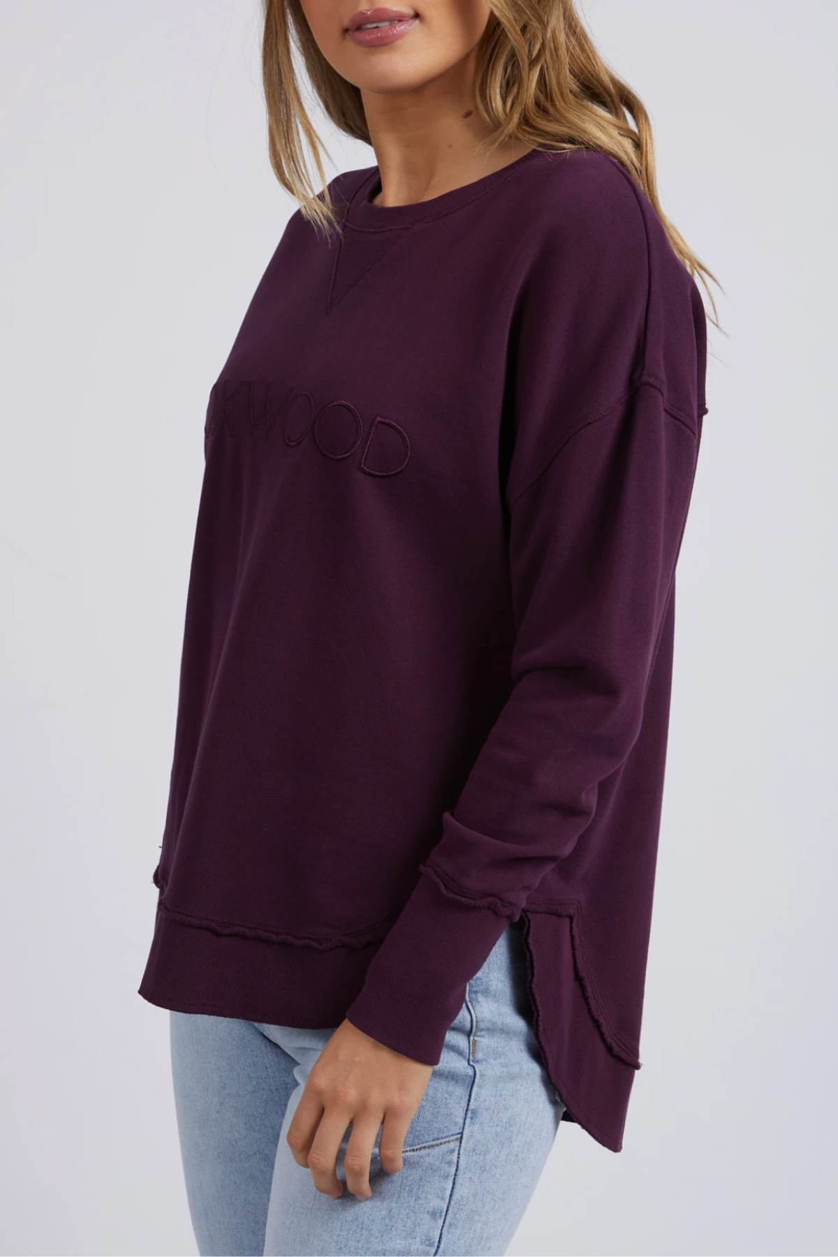 A woman wears a relaxed-fit, long-sleeve Foxwood Simplified Crew sweatshirt in dark purple with subtle embroidery and light blue jeans, standing against a plain light background.