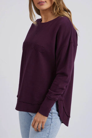 A woman wears a relaxed-fit, long-sleeve Foxwood Simplified Crew sweatshirt in dark purple with subtle embroidery and light blue jeans, standing against a plain light background.