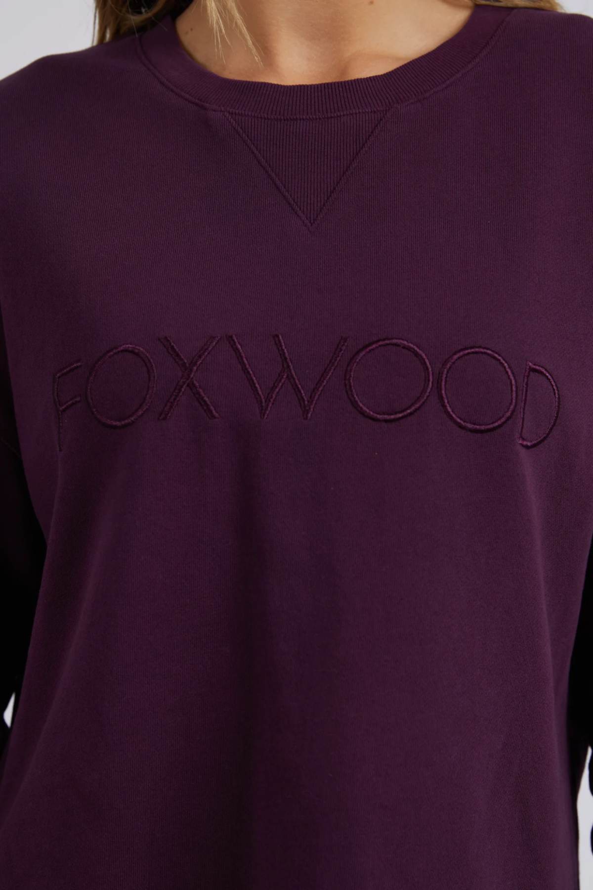 A close-up of a person wearing the Foxwood Simplified Crew, a dark purple 100% cotton sweatshirt with "FOXWOOD" embroidered on the chest. Featuring a relaxed fit for comfort, the person's face is not shown.
