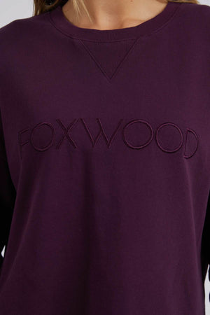 A close-up of a person wearing the Foxwood Simplified Crew, a dark purple 100% cotton sweatshirt with "FOXWOOD" embroidered on the chest. Featuring a relaxed fit for comfort, the person's face is not shown.