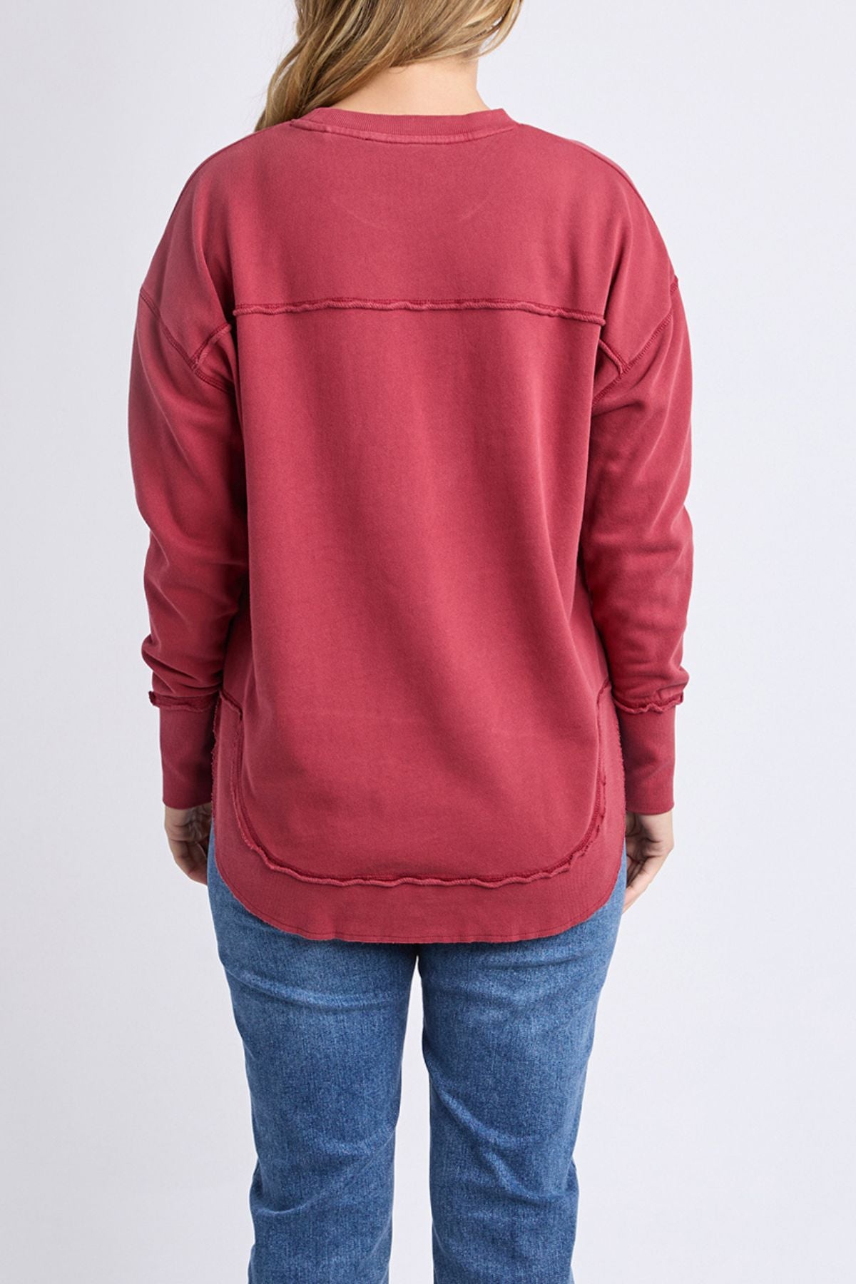 A woman with long, light brown hair is seen from the back wearing a Foxwood Simplified Crew sweatshirt with blue jeans, standing against a plain light background.