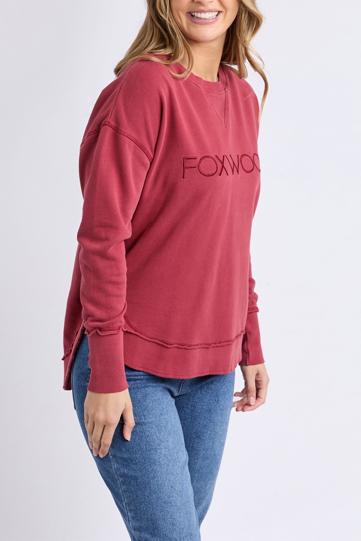 A woman smiles wearing the Foxwood Simplified Crew, a 100% cotton women's relaxed fit sweatshirt in faded red, paired with blue jeans against a light background.