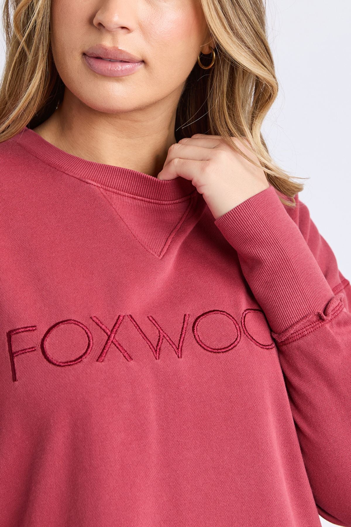 A woman with wavy blonde hair wears the rose-colored Foxwood Simplified Crew, a relaxed fit 100% cotton sweatshirt for women with "FOXWOOD" embroidered on the chest. She touches her collar and wears gold hoop earrings.