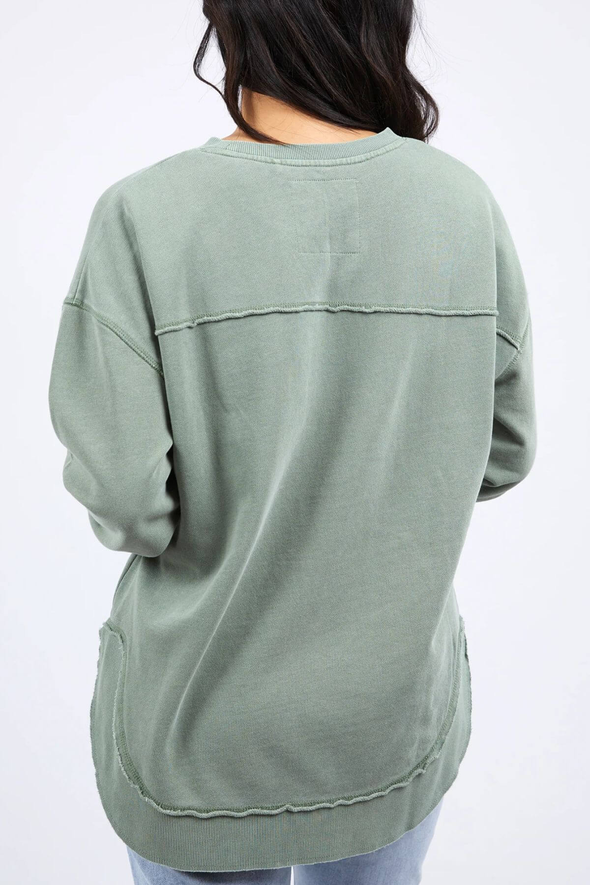 A person with long, dark hair is seen from the back wearing an oversized green sweatshirt with visible stitching details and a relaxed fit, likely the Foxwood Simplified Crew made from 100% cotton. They are also wearing light blue jeans. The background is plain white.