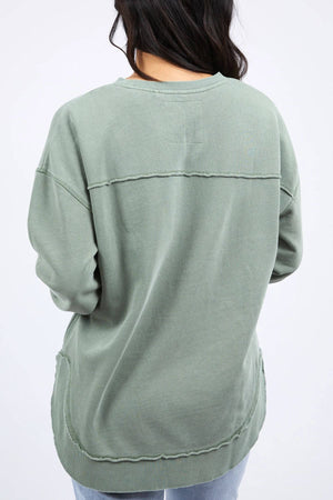 A person with long, dark hair is seen from the back wearing an oversized green sweatshirt with visible stitching details and a relaxed fit, likely the Foxwood Simplified Crew made from 100% cotton. They are also wearing light blue jeans. The background is plain white.