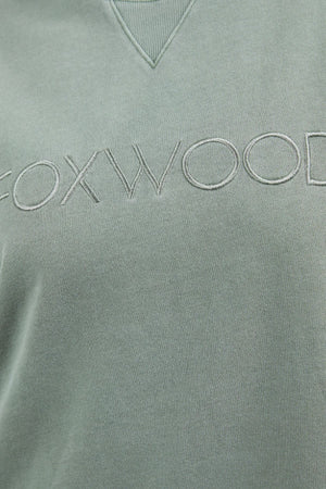 A close-up image of a sage green sweatshirt displaying the text "FOXWOOD" embroidered across the chest in matching green thread. The neckline features a ribbed V-shaped detail. This Foxwood Simplified Crew is made from 100% cotton and offers a relaxed fit for ultimate comfort.