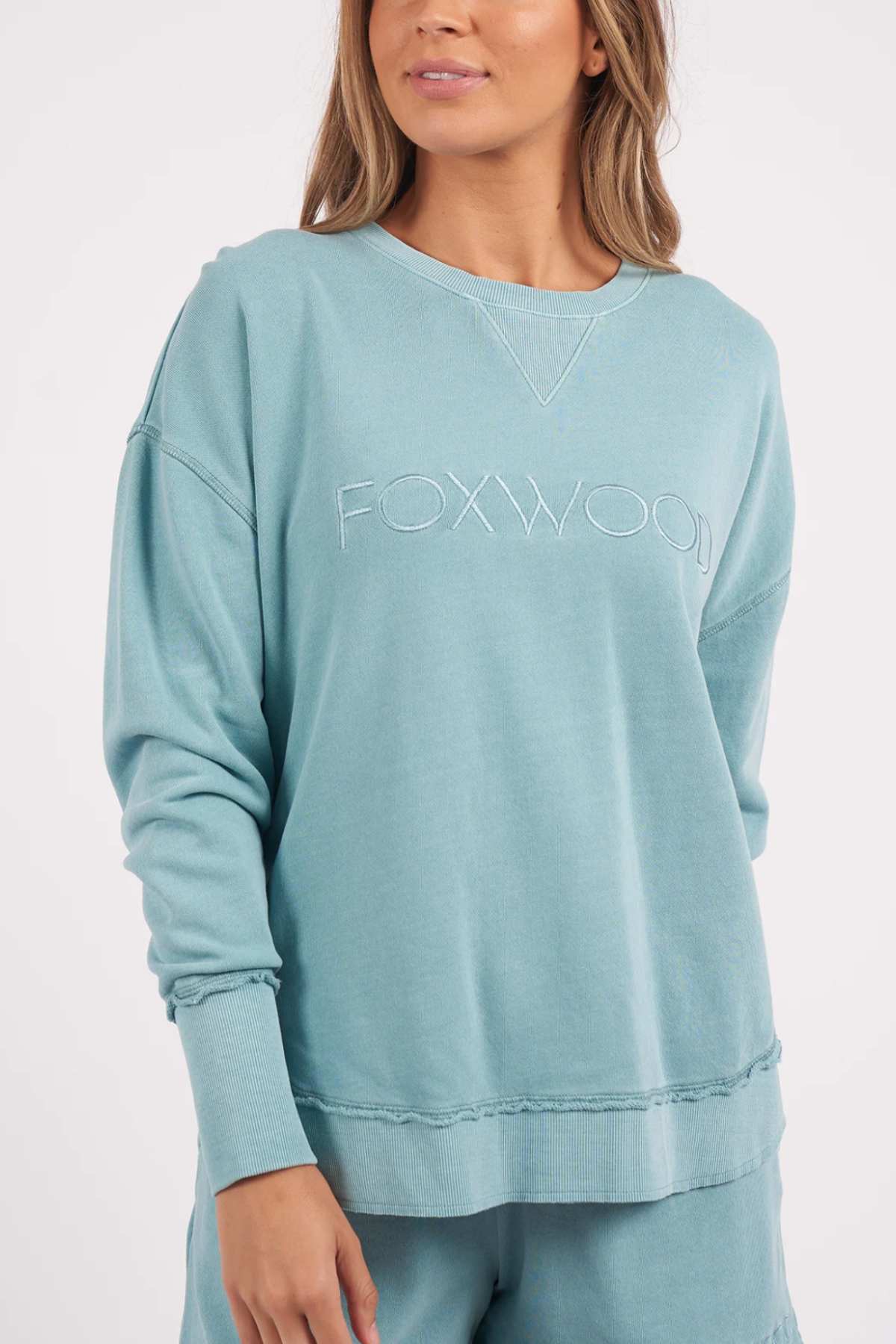 A person wearing the Foxwood Simplified Crew, a light blue sweatshirt with "FOXWOOD" embroidered on the front. The relaxed-fit sweatshirt features a ribbed neckline, cuffs, and hem, along with a V-shaped stitch detail below the collar. The person is also wearing matching pants.