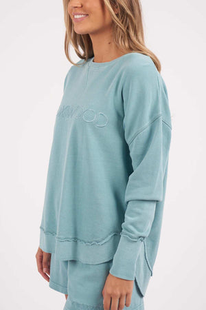 A person is wearing a Foxwood Simplified Crew sweatshirt in teal, featuring the word "KIRKWOOD" embossed on the front. The sweatshirt has a distressed, raw hem detail at the bottom. The person has long, wavy hair and is standing against a plain background.