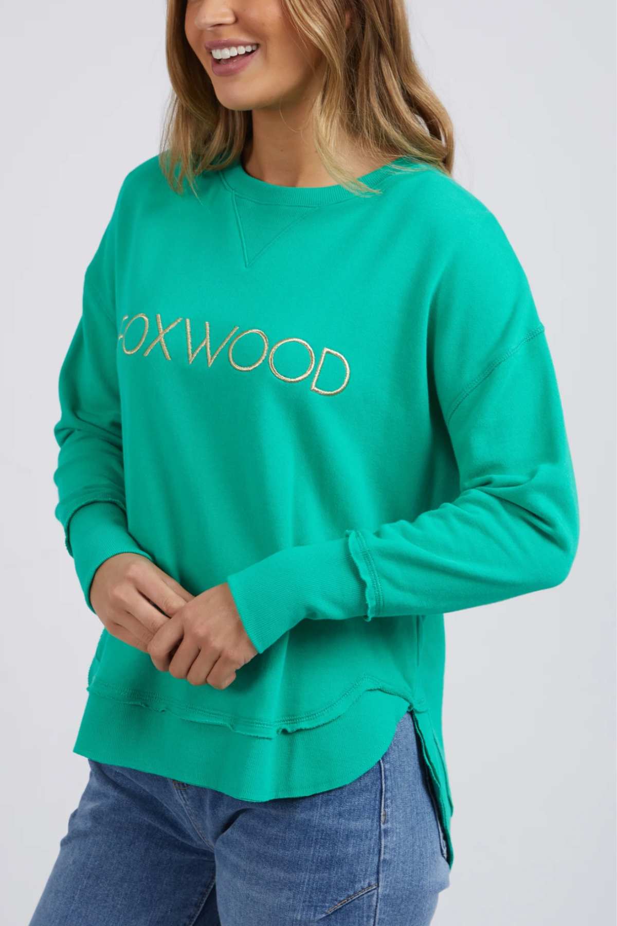 A woman smiles with her hands clasped, wearing the Foxwood Simplified Metallic Crew sweatshirt in bright green with distressed hems and blue jeans.