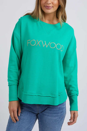 A woman wears the Foxwood Simplified Metallic Crew, a bright green cotton sweatshirt with "FOXWOOD" metallic embroidery across the chest, styled with blue jeans against a plain light background.