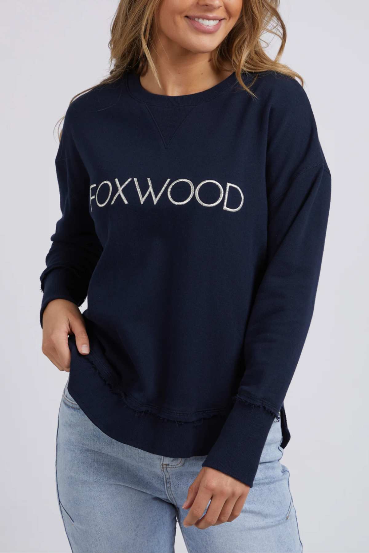 A woman wears the Foxwood Simplified Metallic Crew in navy with white "FOXWOOD" embroidery, paired with light blue jeans. The image is cropped at her shoulders and thighs.