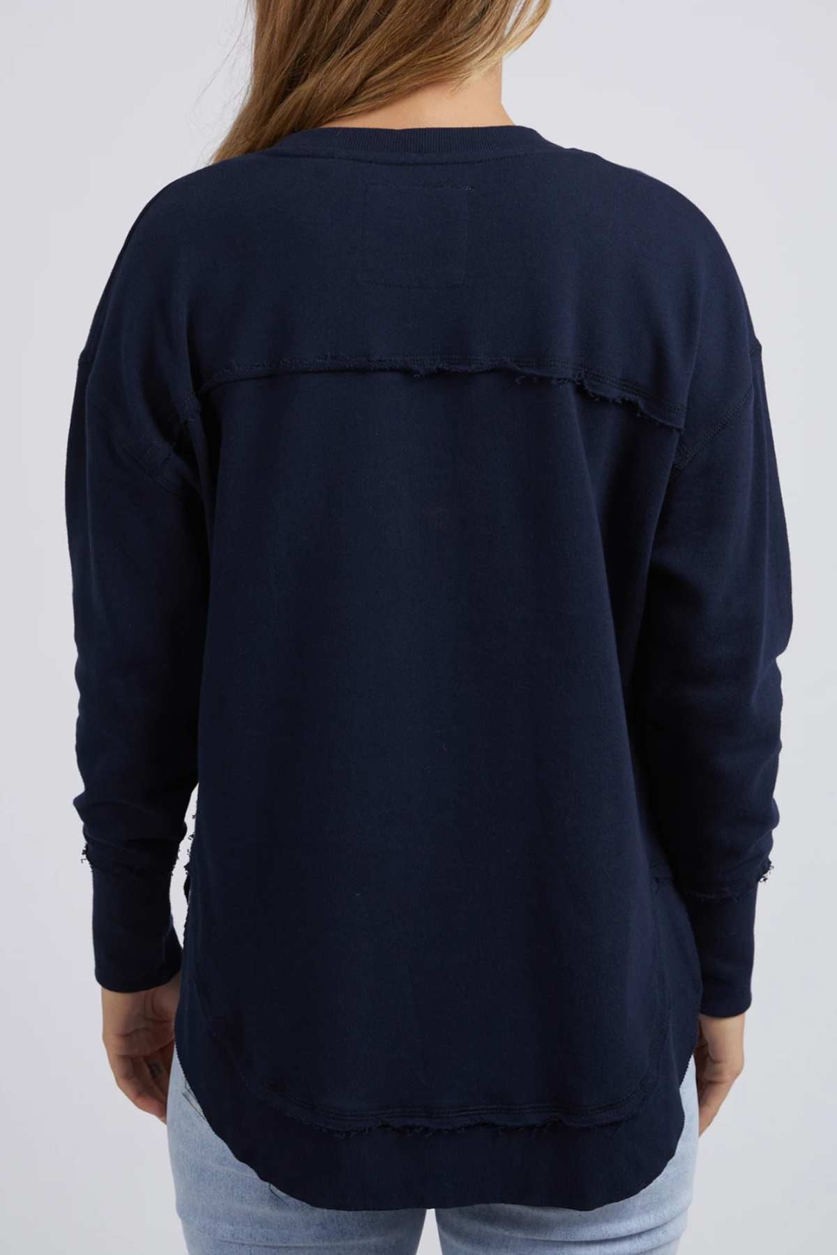 A person with light brown hair wears the Foxwood Simplified Metallic Crew, a navy blue cotton sweatshirt with raw edge seams and chest embroidery, paired with light blue jeans against a plain, light-colored background.