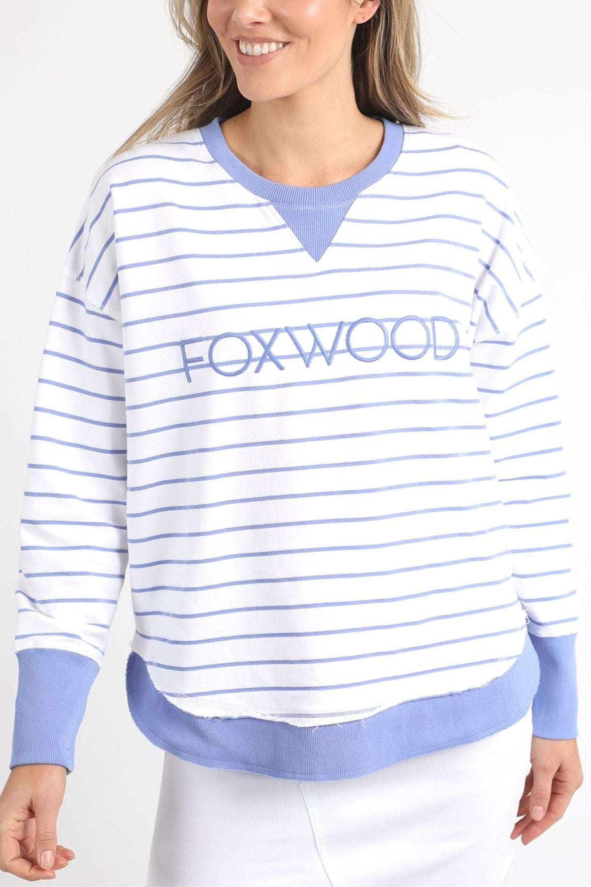 The individual is dressed in the Foxwood Simplified Stripe Crew, a white and light blue striped sweatshirt made entirely of cotton, featuring "FOXWOOD" embroidered boldly on the front. The cuffs and collar are a solid light blue color. They pose against a simple backdrop with a slight smile.
