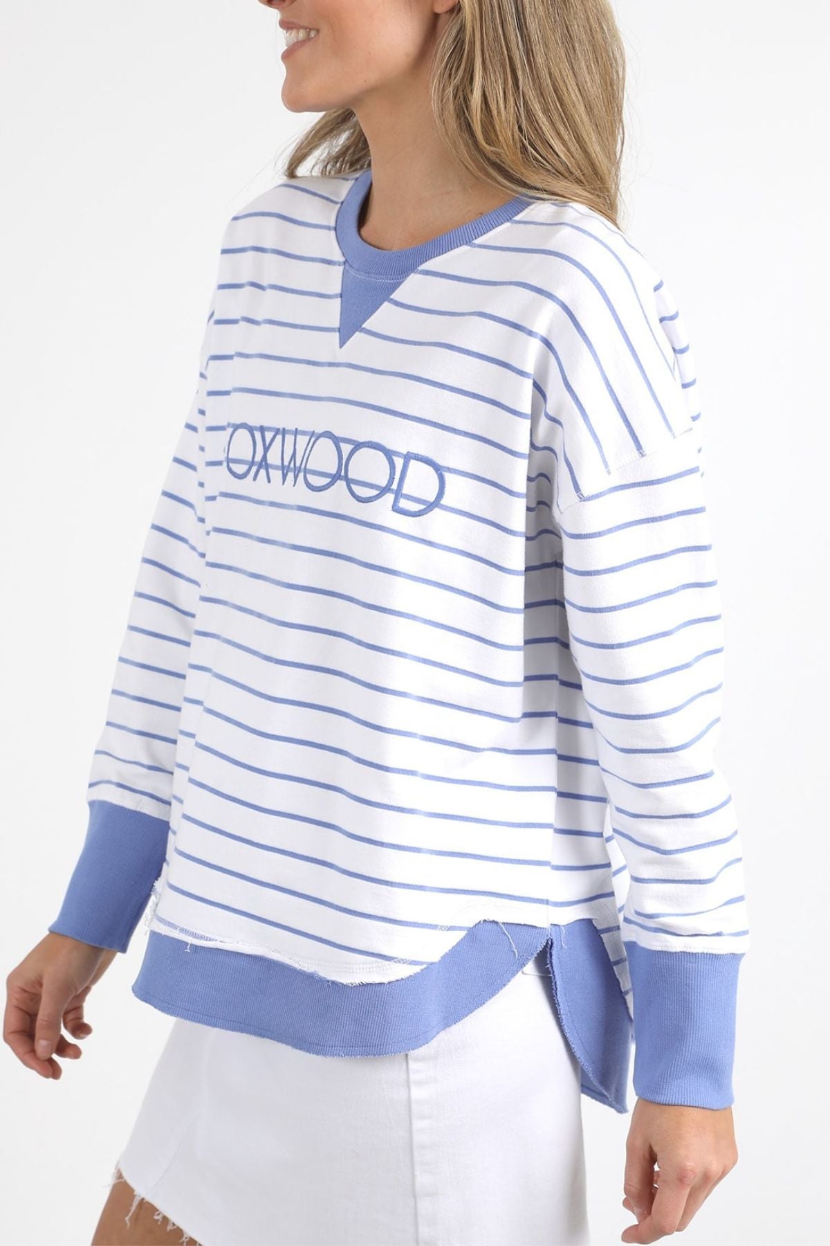 A woman wears the Foxwood Simplified Stripe Crew, featuring white and light blue stripes with "OXWOOD" embroidered on the front. Crafted from 100% cotton, it is designed with blue ribbed cuffs and hem. She is set against a plain white background, highlighting its classic style.