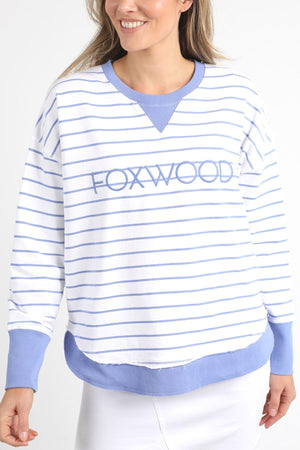 The individual is dressed in the Foxwood Simplified Stripe Crew, a white and light blue striped sweatshirt made entirely of cotton, featuring "FOXWOOD" embroidered boldly on the front. The cuffs and collar are a solid light blue color. They pose against a simple backdrop with a slight smile.