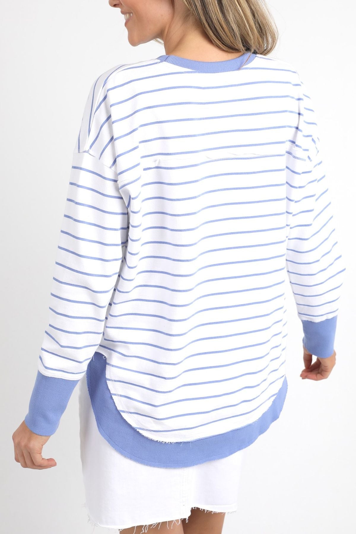 A person wearing the Foxwood Simplified Stripe Crew, featuring a 100% cotton white design with blue stripes and cuffs, stands with their back to the camera. They pair it with a white skirt, embodying timeless style against a plain white background.