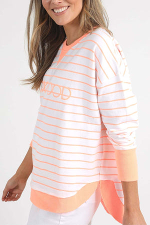A person is smiling while wearing the Foxwood Simplified Stripe Crew, a relaxed-fit, crew neck white and peach striped sweater featuring "ROAD" and subtle Foxwood embroidery. The sweater has a high-low hem and peach cuffs, paired with white pants. They're standing against a plain white background, exuding effortless style.
