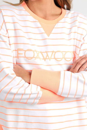 A person is wearing the Foxwood Simplified Stripe Crew, a crew neck long-sleeve shirt adorned with orange and white stripes. The word "FOXWOOD" is elegantly embroidered across the chest. This 100% cotton shirt features crossed arms and boasts an orange neckline and cuffs for a stylish finish.