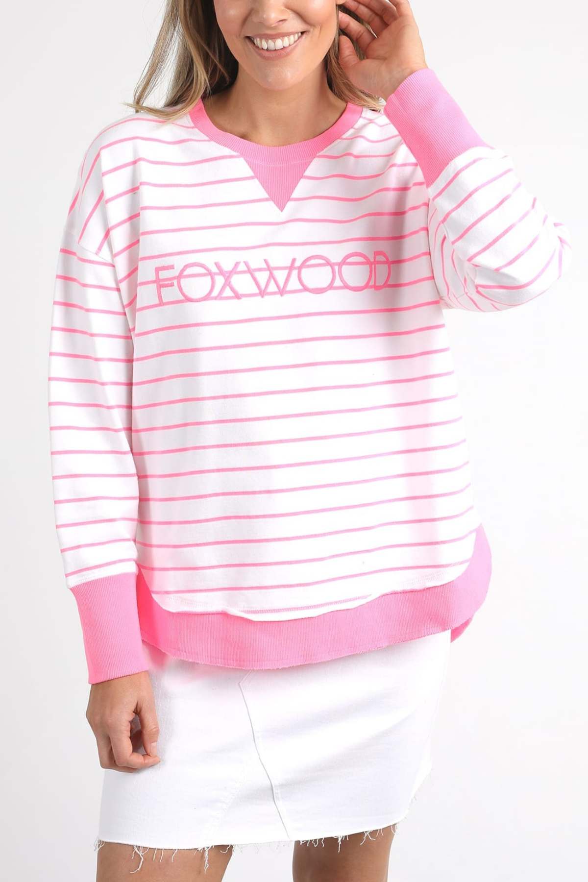 A person is wearing a Foxwood Simplified Stripe Crew in white and pink stripes, known for its relaxed fit and made from 100% cotton, paired with a white skirt. They are smiling with a hand near their hair against a plain white background.