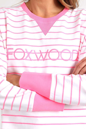 A person dressed in the Foxwood Simplified Stripe Crew, featuring white and pink stripes and crafted entirely from 100% cotton, stands with arms crossed. The word "FOXWOOD" is prominently displayed across the chest. This relaxed fit sweater is completed with a solid pink neckline and cuffs, blending comfort with style seamlessly.
