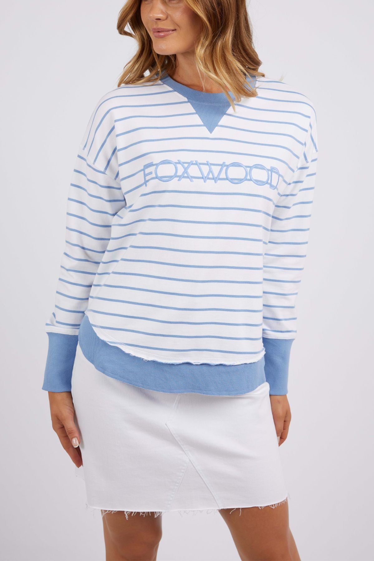A woman poses in front of a simple backdrop, her wavy hair falling over a Foxwood Simplified Stripe Crew sweatshirt designed with light blue and white yarn-dye stripes. She elegantly pairs it with a white skirt, capturing the charming essence of Foxwood Clothing.
