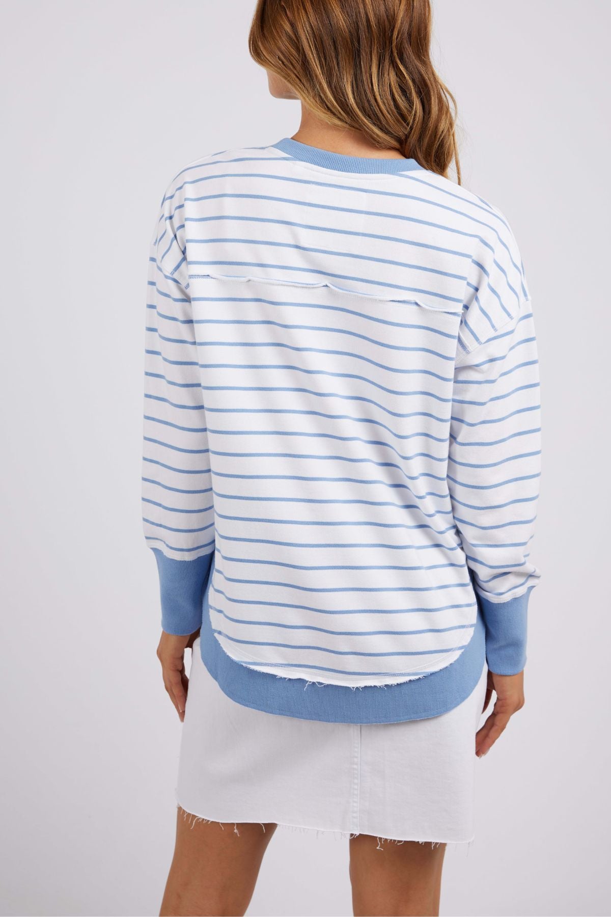 A person with long hair is seen from behind wearing the Foxwood Simplified Stripe Crew, a white and light blue striped sweater with blue cuffs and hem, along with a white skirt. The outfit is completed by the above-the-knee skirt featuring a stylish frayed hem for added flair.